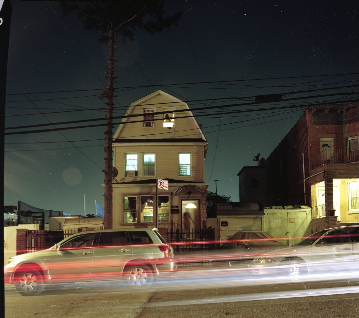 Neighborhood street at night with cars passing by, featuring a two-story house.